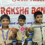 Three smiling schoolboys in beige uniforms pose with bright decorative medals and ribbons in front of a large banner